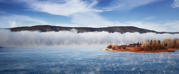 Winter frosty fog on an unfrozen river. Trees and grass on the banks.