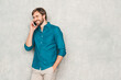 © halayalex - Portrait of handsome smiling hipster lumbersexual businessman model wearing casual jeans shirt clothes. Fashion stylish man posing against gray wall in studio in eyewear. Talking on the smartphone
