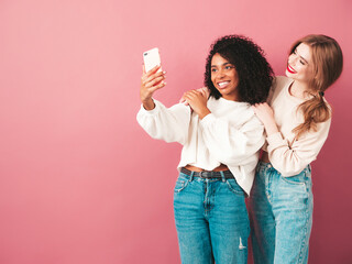 Two young beautiful smiling international hipster female in trendy summer jeans clothes. Sexy carefree women posing near pink wall in studio. Positive models having fun. They taking selfie photos