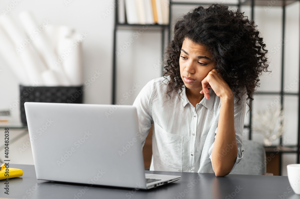 Young African American female trainee or an office worker with Afro hairstyle sitting at the desk, feeling bored and sleepy does not want to do homework or work on the project, unhappy with the task - obrazy, fototapety, plakaty 