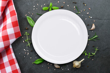 Classic Italian cooking template - top view of white blank ceramic plate on vintage black stone kitchen countertop with  red checkered tablecloth and selection of herbs and spices.