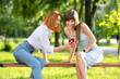 © bilanol - Two young women friends sitting on a bench in summer park looking in smartphone.