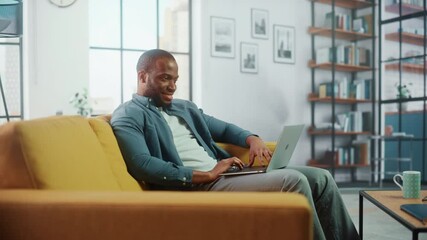 Wall Mural - Handsome Black African American Man Working on Laptop Computer while Sitting on a Sofa in Cozy Living Room. Freelancer Working From Home. Browsing Internet, Using Social Networks, Having Fun in Flat.