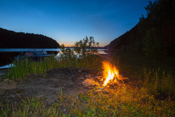 Wall Mural - A large night fire on the river bank in a forest clearing, flames, sparks, blue sky, light beyond the horizon, a boat near the shore.