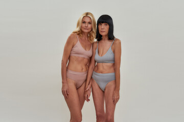 Two attractive caucasian women in underwear looking at camera, holding hands while posing together over light background. Warmth in relation, mature beauty concept