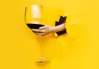 A left woman's hand emerges through a torn hole in yellow paper with a large glass of red wine. The concept of alcoholism, drunkenness and hangover. Copy space, wineglass.