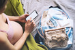 Pregnant woman getting ready for labor packing stuff for hospital, making notes or checklist in smartphone. Baby clothes, necessities for mother and newborn in maternity bag.