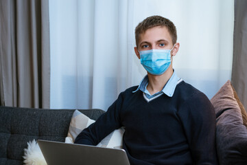 young man sitting on sofa with laptop in medical mask