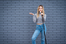 Young Blonde Female Comedian Against Blue Grey Brick Wall With Microphone And Expressions