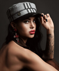 Portrait of young brunette woman with pouty lips, tattoo and piercing sitting sideways holding her cap visor and looking at camera over dark background. Stylish look, fashion, glamour concept