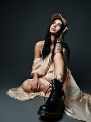 Sexy brunette woman with tattoo on arm and piercing on face in cap, evening maxi dress and massive brutal shoes sits on floor looking up over black background. Stylish look, fashion, glamour concept