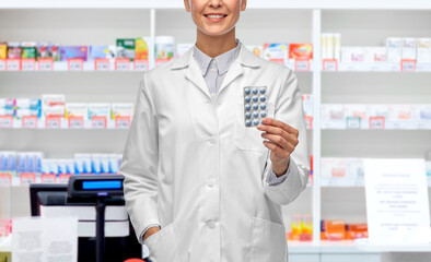 Canvas Print - medicine, profession and healthcare concept - happy smiling female doctor in white coat holding pills over pharmacy background