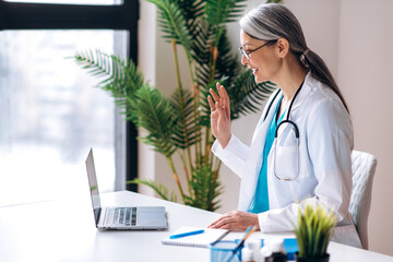 Female practitioner middle aged doctor wearing a lab coat and headset speaking by video call using laptop with unhealthy patient, online consultation. Remote medical help concept