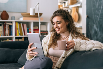 woman relaxed sitting on sofa in her home and drinking morning coffee and using digital tablet