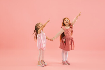 Pointing. Childhood and dream about big and famous future. Pretty little girls on coral pink studio background. Childhood, dreams, imagination, education, facial expression, emotions concept.