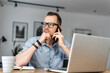 © Vadim Pastuh - A busy young entrepreneur wearing glasses and smart casual shirt is talking on the mobile phone and using a laptop. Remote work concept