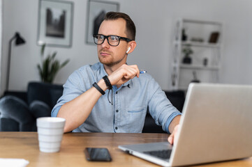 An intelligent young man in glasses and casual shirt sits at the desk with a laptop and looks away lost in thoughts. A freelancer is thinking about solving business tasks