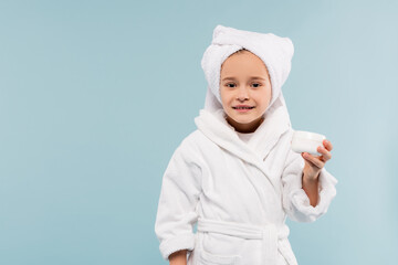 smiling child in bathrobe and towel on head holding container with cosmetic product isolated on blue