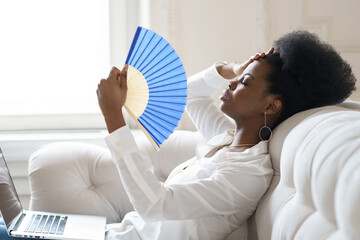 Tired Afro business woman suffering from heatstroke or hot summer flat without air-conditioner, touching her forehead, using waving fan, working on laptop, sitting in living room at home. Overheating