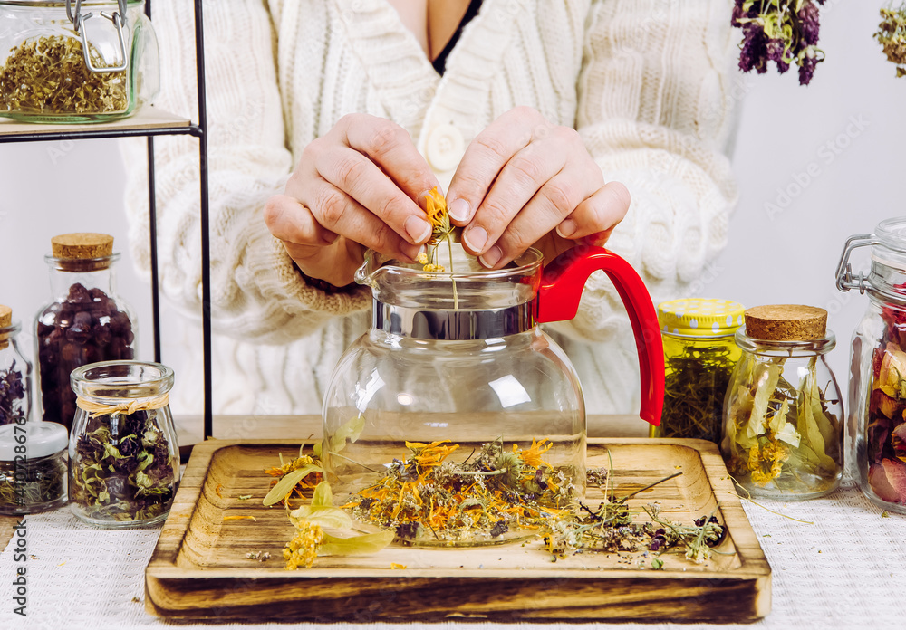 Close up view of woman herbalist mixing various dried herbs for traditional medicinal tea. Dried herbs in glass jars on background. - obrazy, fototapety, plakaty 
