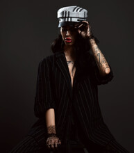 Young Woman With Pouty Lips And Tattoo On Body Arms In Pinstripe Pantsuit On Naked Body And Gloves Sits Holding Visor Of Her Cap Over Dark Background. Stylish Look, Fashion, Glamour Concept