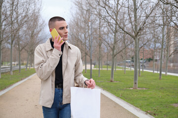 Wall Mural - Handsome young man wearing make up, walking and speaking on his smartphone, carrying shopping bags. Non binary androgynous guy.