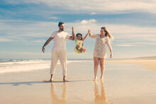 Cheerful Parents And Little Girl Enjoying Walking And Activities On Beach, Kid Holding Parents Hands, Jumping And Throwing Legs Up. Front View. Family Outdoor Activities Concept