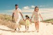 © Mangostar - Happy family couple and little kid in summer clothes walking white along sand path, girl holding parents hands. Front view. Family outdoors concept