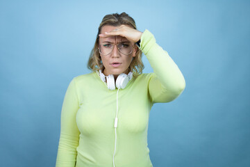 Young caucasian woman wearing headphones on neck over blue background very happy and smiling looking far away with hand over head. searching concept.