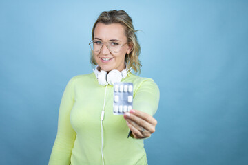 Wall Mural - Young caucasian woman wearing headphones on neck over blue background holding tablet of pills medicines looking positive and happy standing and smiling with a confident smile showing teeth