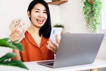 Happy Asian Woman Successful Make Money Online Hand Holding Thai Baht Money And Smart Phone With Laptop On Desk ,e-business Concept