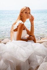 Wall Mural - Beautiful sexy blonde woman in wedding dress with long hair and tanned body sitting at the seashore.