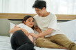 © ake - Asian pregnant woman and boyfriend looking at clothes for new born baby on bed at home