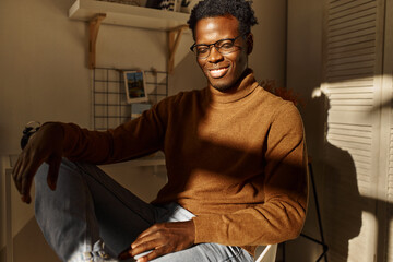 Wall Mural - Horizontal image of joyful successful young African American businessman working from home in stylish sunlit room, smiling happily with his eyes closed, sitting comfortably in relaxed posture