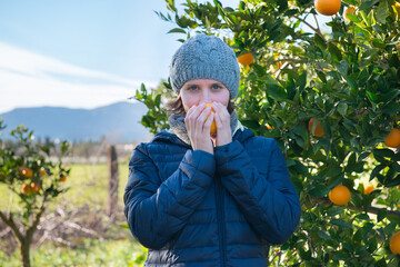 Portrait of a beautiful blue-eyed young woman wearing a cap smelling an orange next to an orange tree