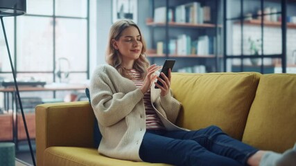 Wall Mural - Beautiful Caucasian Female Using Smartphone in Stylish Living Room while Resting on a Cozy Couch Sofa. Young Woman at Home, Browsing Internet, Using Social Networks, Having Fun in Flat.