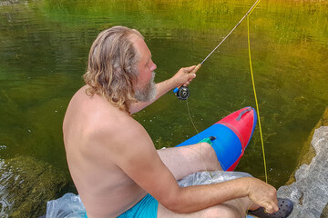 Mature gray-haired bearded man is sitting with a rod on the lake in nature and fishing. Summer, sunlight. Rest, relaxation concept.