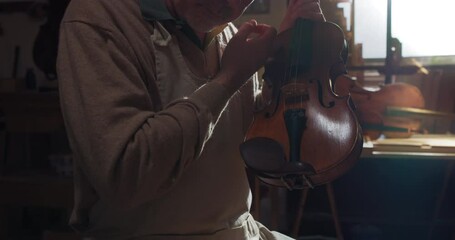 Poster - Cinematic shot of experienced master artisan luthier controlling quality of handmade wood violin just made in creative workshop. Concept of spiritual instrument, art, orchestra, passion for music.