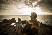 Digital Nomad People At Work Outdoor With Computer Laptop And 5g Internet Connection In Roaming Phone Hotspot - Man Travel And Work Alone Sitting And Enjoying The Sunset On The Ocean Coast