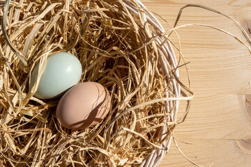 Organic Easter concept: Top view on a basket filled with straw and two natural colored eggs from the farm. Sun light falling on still life. Wood table surface in background with copy space