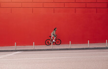 Sporty Woman In Casual Clothes Rides A Bicycle On A City Street On A Background Of A Red Wall And Looks At The Camera. Weekend Cycling As A Sport