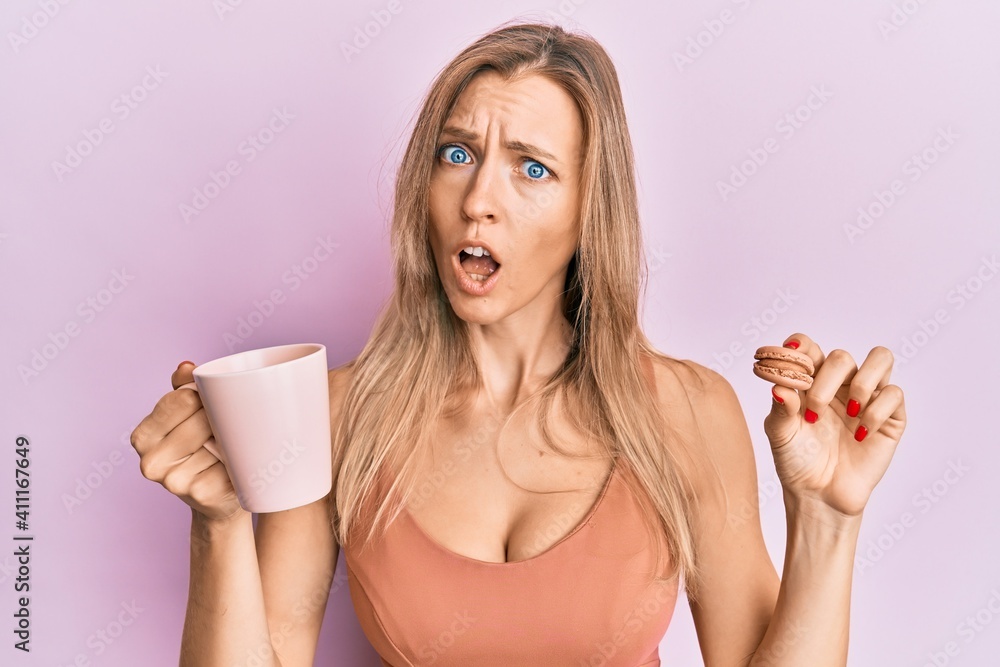 Beautiful caucasian woman drinking coffee and eating french pastry macaron in shock face, looking skeptical and sarcastic, surprised with open mouth - obrazy, fototapety, plakaty 