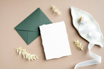 Wall Mural - Wedding stationery top view. Flat lay blank white invitation card, green envelope, wedding decorations, silk ribbon and dried flowers on pastel beige background.