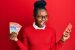 Young african american woman using smartphone holding swiss franc banknotes angry and mad screaming frustrated and furious, shouting with anger. rage and aggressive concept.