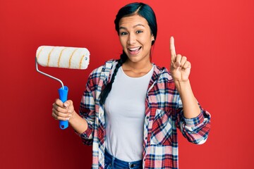 Wall Mural - Beautiful hispanic woman holding roller painter smiling with an idea or question pointing finger with happy face, number one