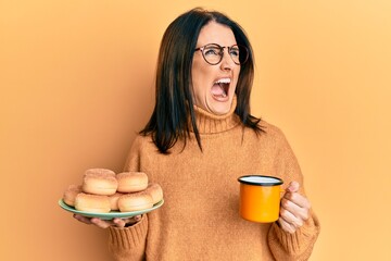 Poster - Middle age brunette woman eating doughnuts and drinking a cup of coffee angry and mad screaming frustrated and furious, shouting with anger. rage and aggressive concept.