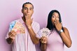 Young latin couple holding mexican pesos covering mouth with hand, shocked and afraid for mistake. surprised expression