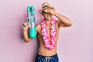 Poster - Young caucasian man wearing swimsuit and hawaiian lei holding watergun stressed and frustrated with hand on head, surprised and angry face