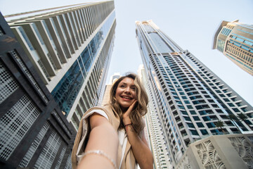 Wall Mural - Young woman take selfie photo on the phone with modern skycrapers on background. Low angle