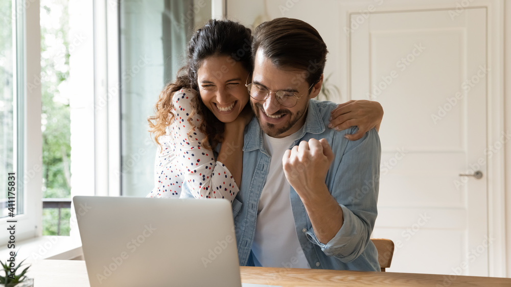 Laughing excited millennial family couple embracing by computer screen winning prize at lottery or sports betting. Loving young wife sharing amazed husband joy of getting reward perfect job proposal - obrazy, fototapety, plakaty 